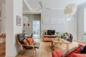 Ein modernes Wohnzimmer mit Sofa, Sessel, Couchtisch und Fernseher. Eine Deckenventilator, Kunstwerke an den Wänden und eine Tür zu einem weiteren Raum sind vorhanden.