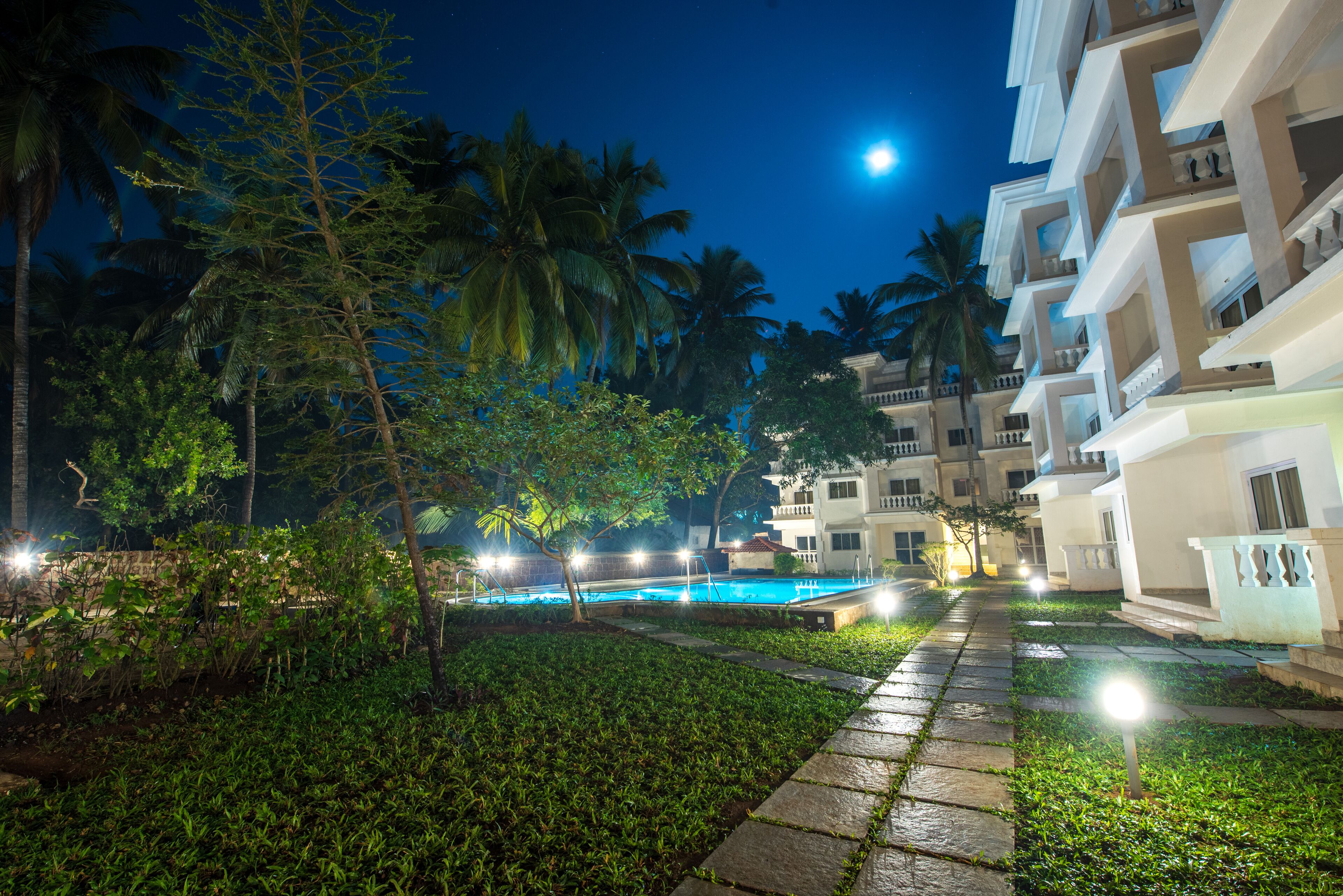 A nighttime resort scene with a swimming pool, palm trees, and multi-story buildings.