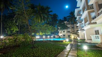 A nighttime resort scene with a swimming pool, palm trees, and multi-story buildings.