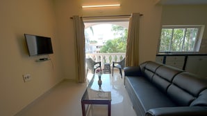 Una sala de estar con sofá, mesa de centro, televisor y balcón con vista a edificios y árboles.