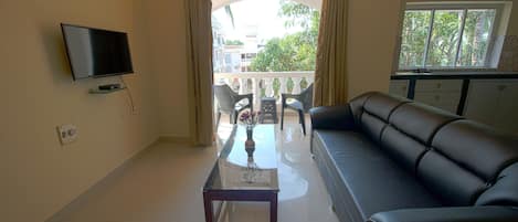 Una sala de estar con sofá, mesa de centro, televisor y balcón con vista a edificios y árboles.