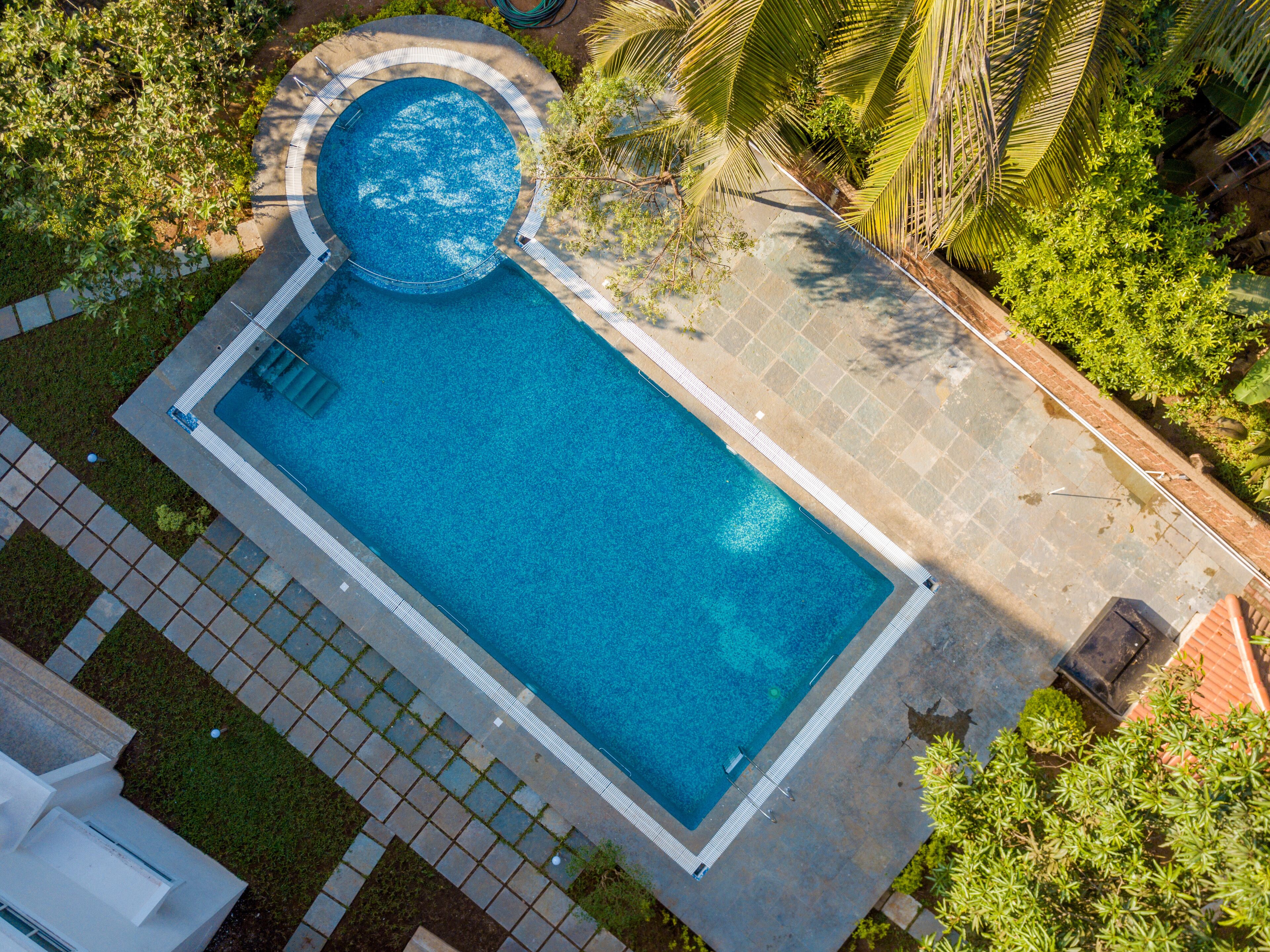 A rectangular swimming pool with a circular hot tub area, surrounded by a stone deck and lush greenery.