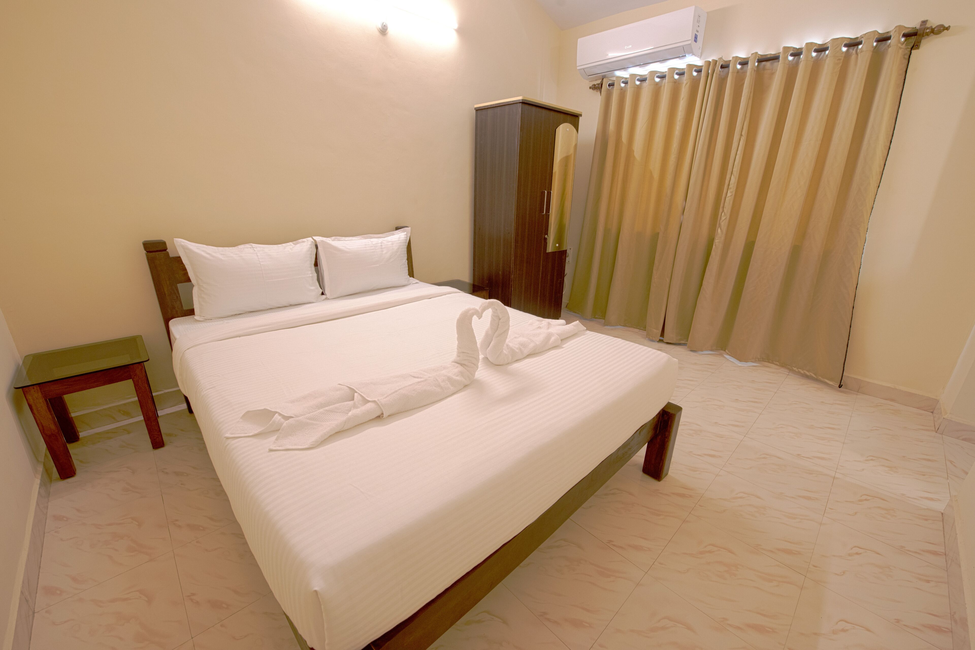 A neatly made bed with white linens, a wooden headboard, a small wooden nightstand, and a wardrobe in a room with beige walls and tiled flooring.