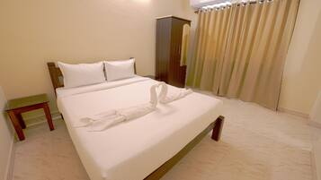 A neatly made bed with white linens, a wooden headboard, a small wooden nightstand, and a wardrobe in a room with beige walls and tiled flooring.