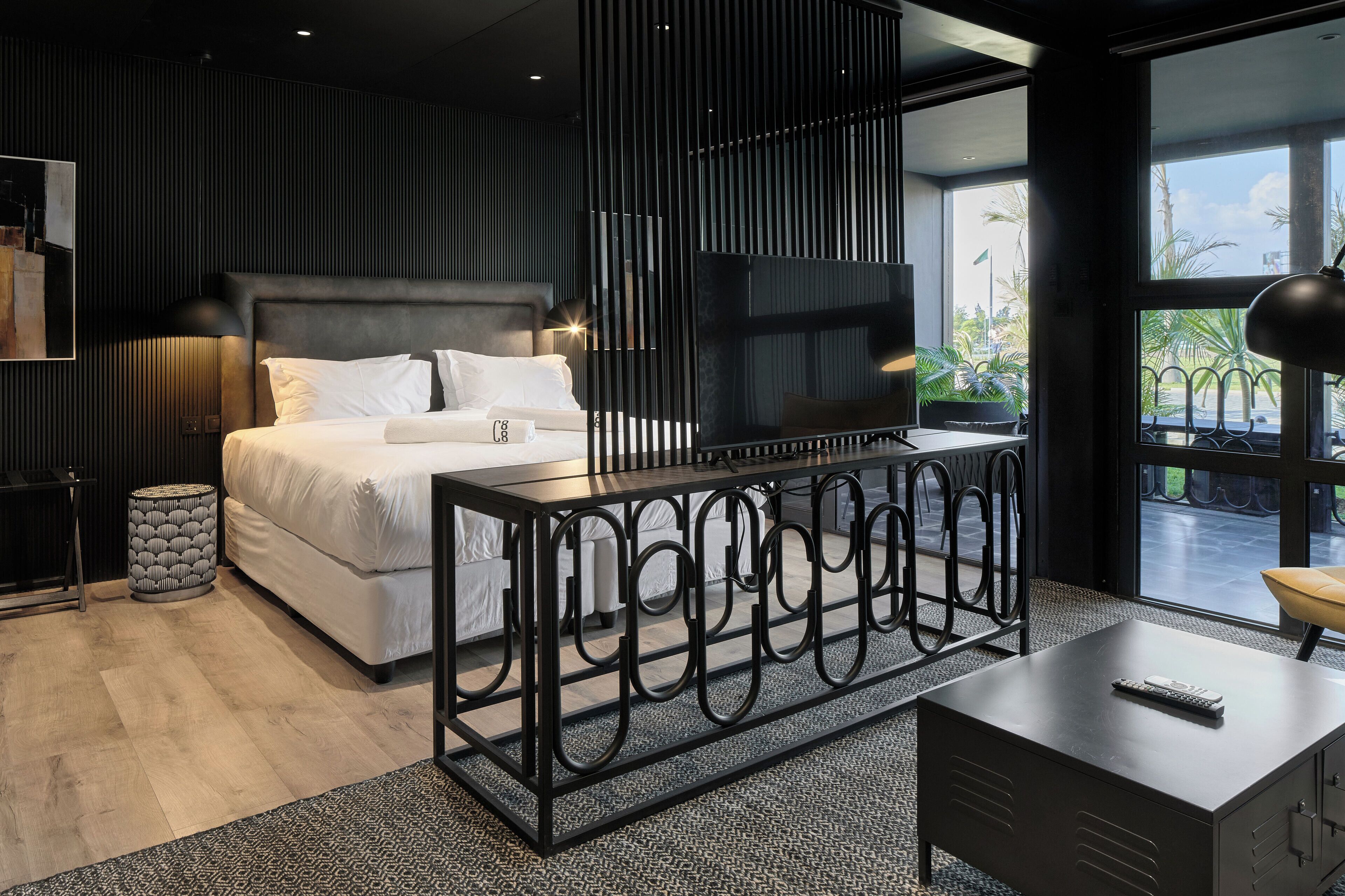 A modern bedroom with a large bed, a TV, a desk, and a view of palm trees outside.