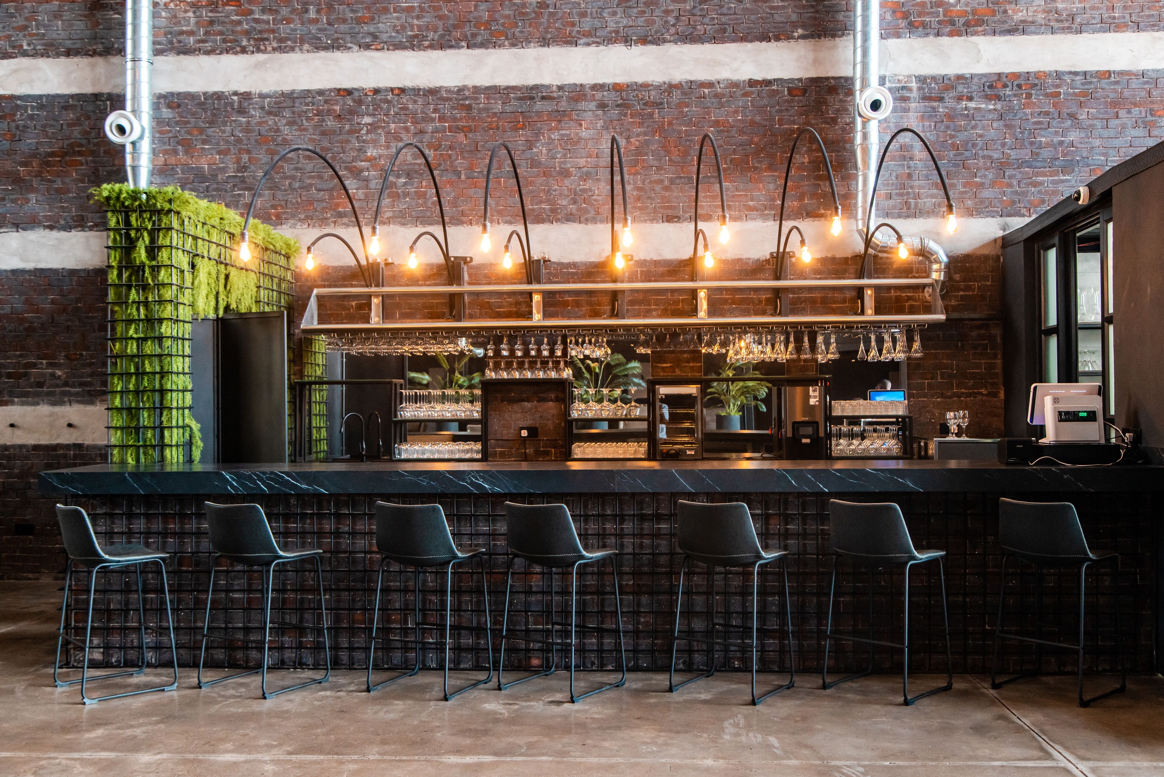 Een bar met een bakstenen muur, groene planten en barstoelen.