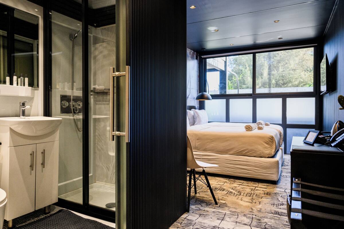 A modern hotel room with a glass shower enclosure, a bed with neatly rolled towels, a desk with a telephone, and a large window offering a view of the outdoors.