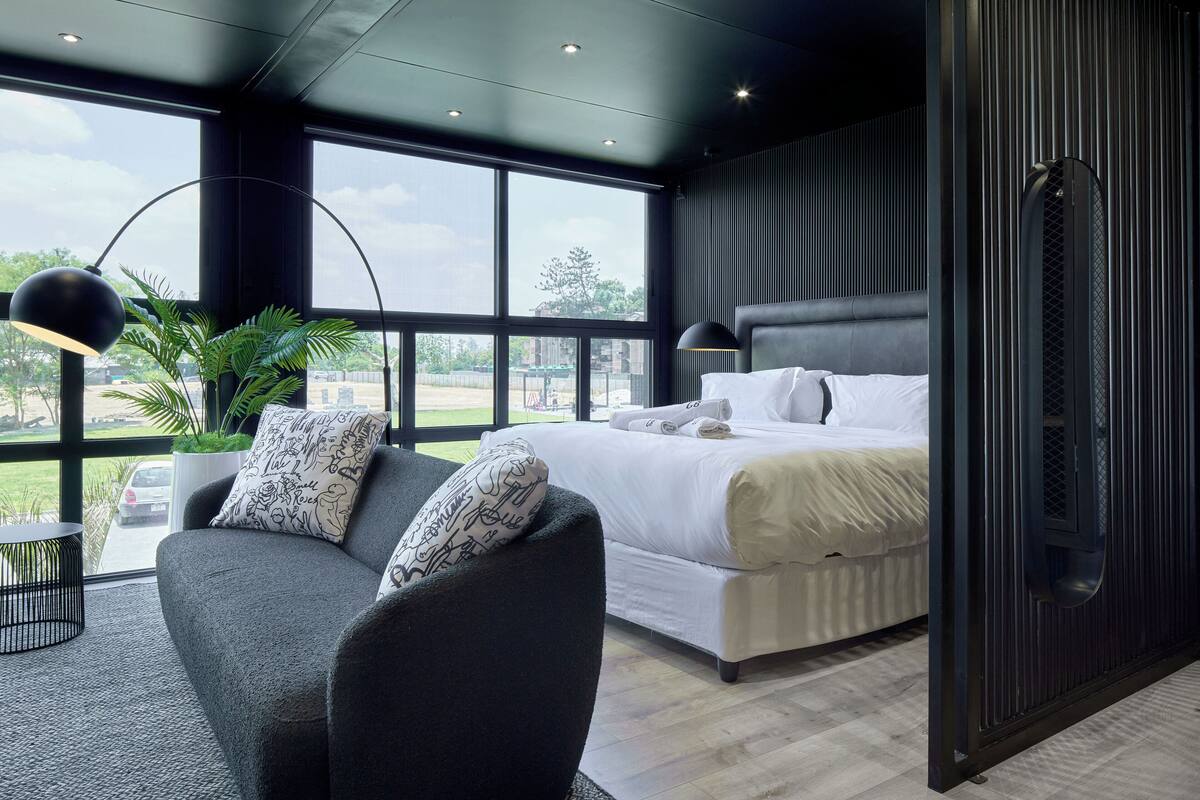 A modern bedroom with a large bed, a dark upholstered chair, a potted plant, and a view of the outdoors.