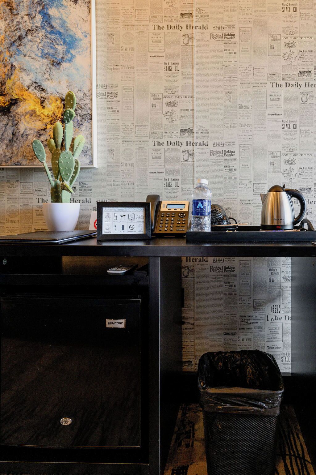A desk with a cactus, a telephone, a water bottle, and a kettle, with newspapers as a backdrop.