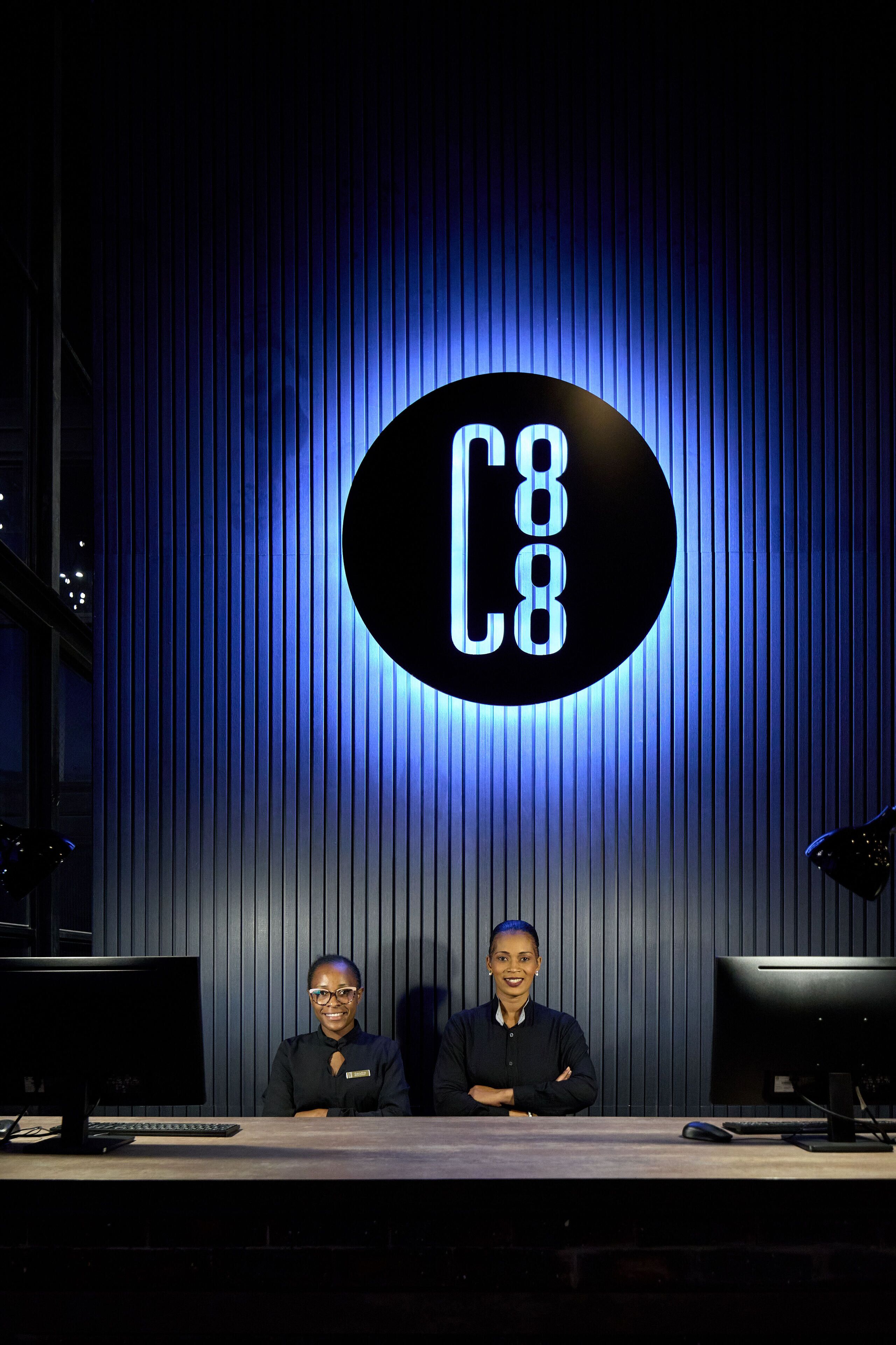 Two individuals seated at a desk with a computer and keyboard, in front of a wall with vertical lines and a circular sign with the letters "C8" illuminated in blue.