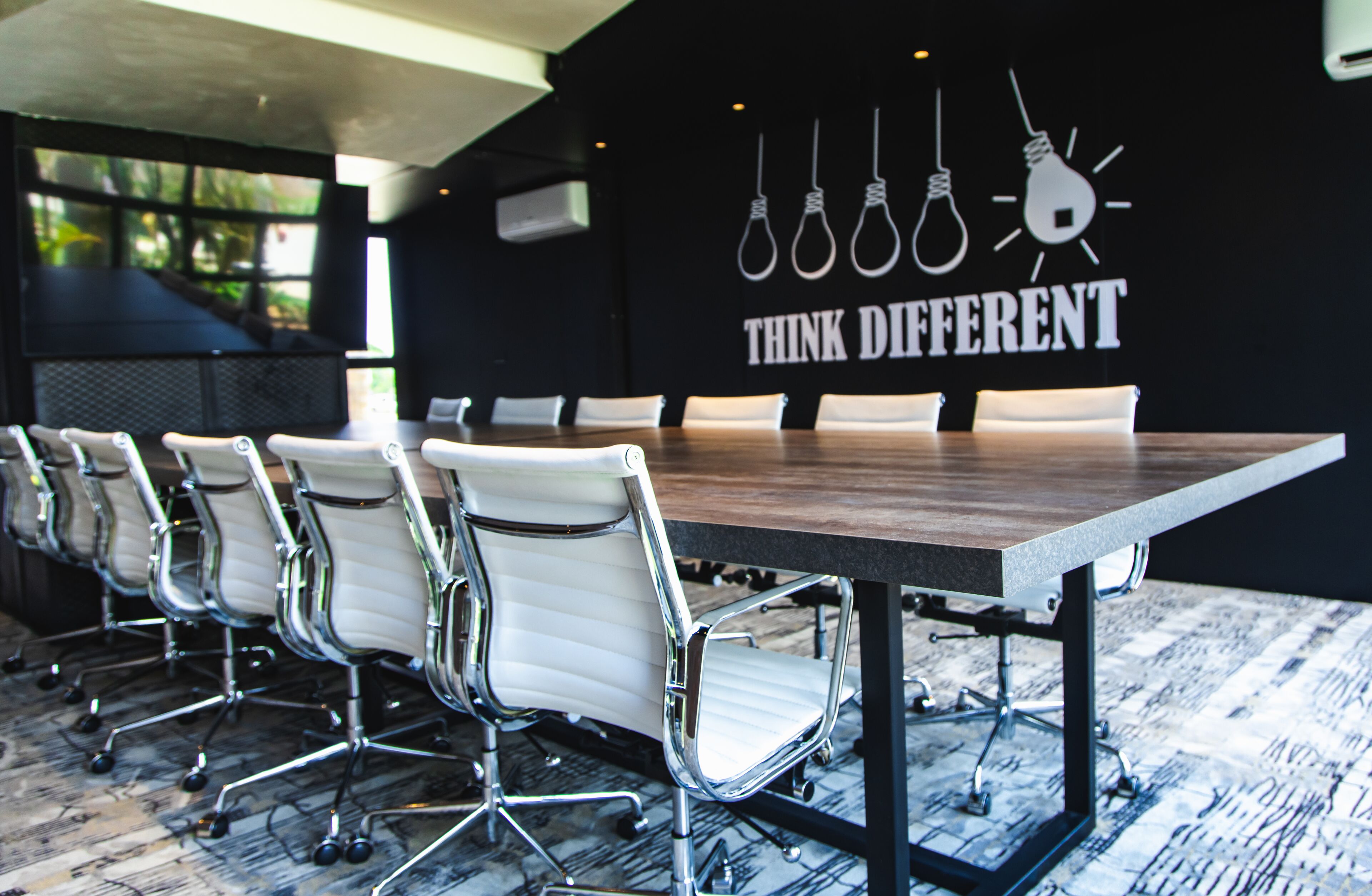 A conference room with a long table and white chairs, a black wall with a 'Think Different' poster, and a large window with a view of greenery.