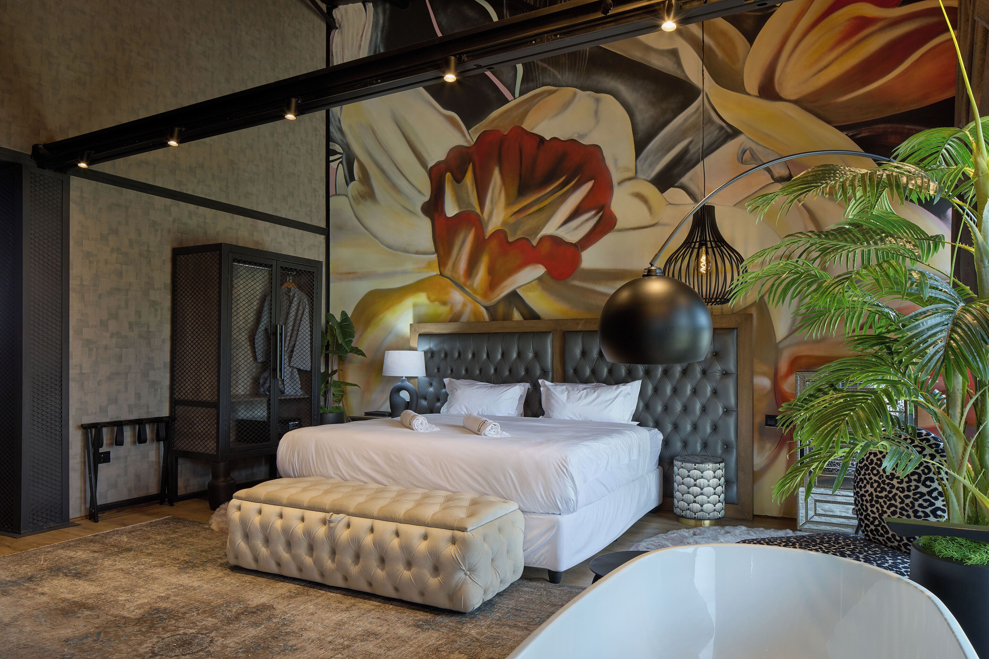 Un dormitorio moderno con una cama grande, un otoman tapizado, una planta grande en maceta y un mural original en la pared.