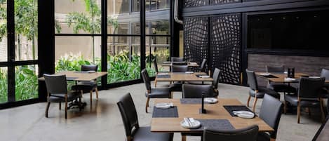 A modern restaurant with large windows, indoor plants, and a mix of wooden and black chairs.