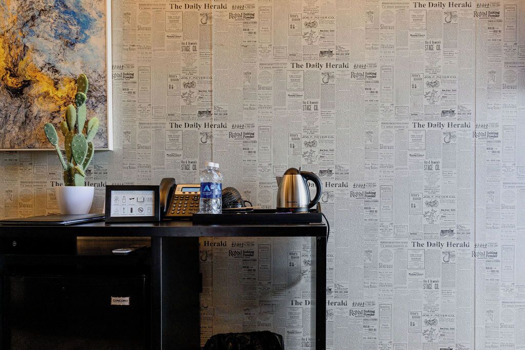 A desk with a cactus, a framed picture, a telephone, a water bottle, and a coffee pot against a wall covered with newspaper clippings.