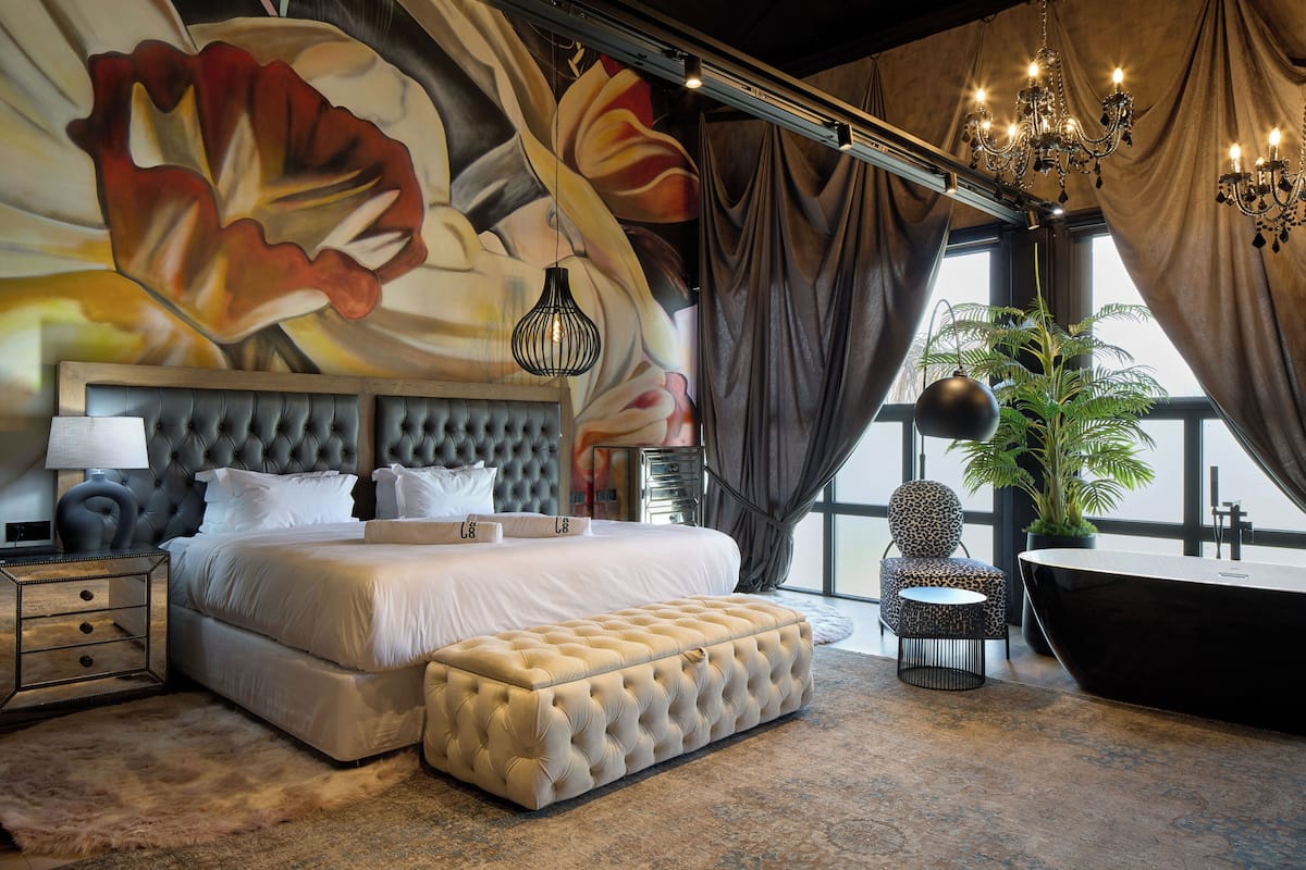 A luxurious bedroom with a large bed, a tufted bench, a round side table, a large potted plant, a freestanding bathtub, and a mural of flowers on the wall.