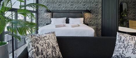 Un dormitorio moderno con una cama grande, un sofá oscuro y un ventanal amplio.