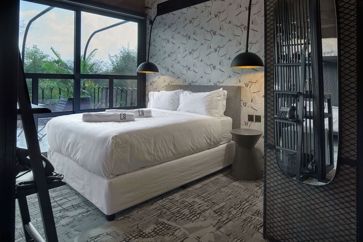 A bedroom with a large bed, two bedside lamps, a window with a view of greenery, and a patterned wall.