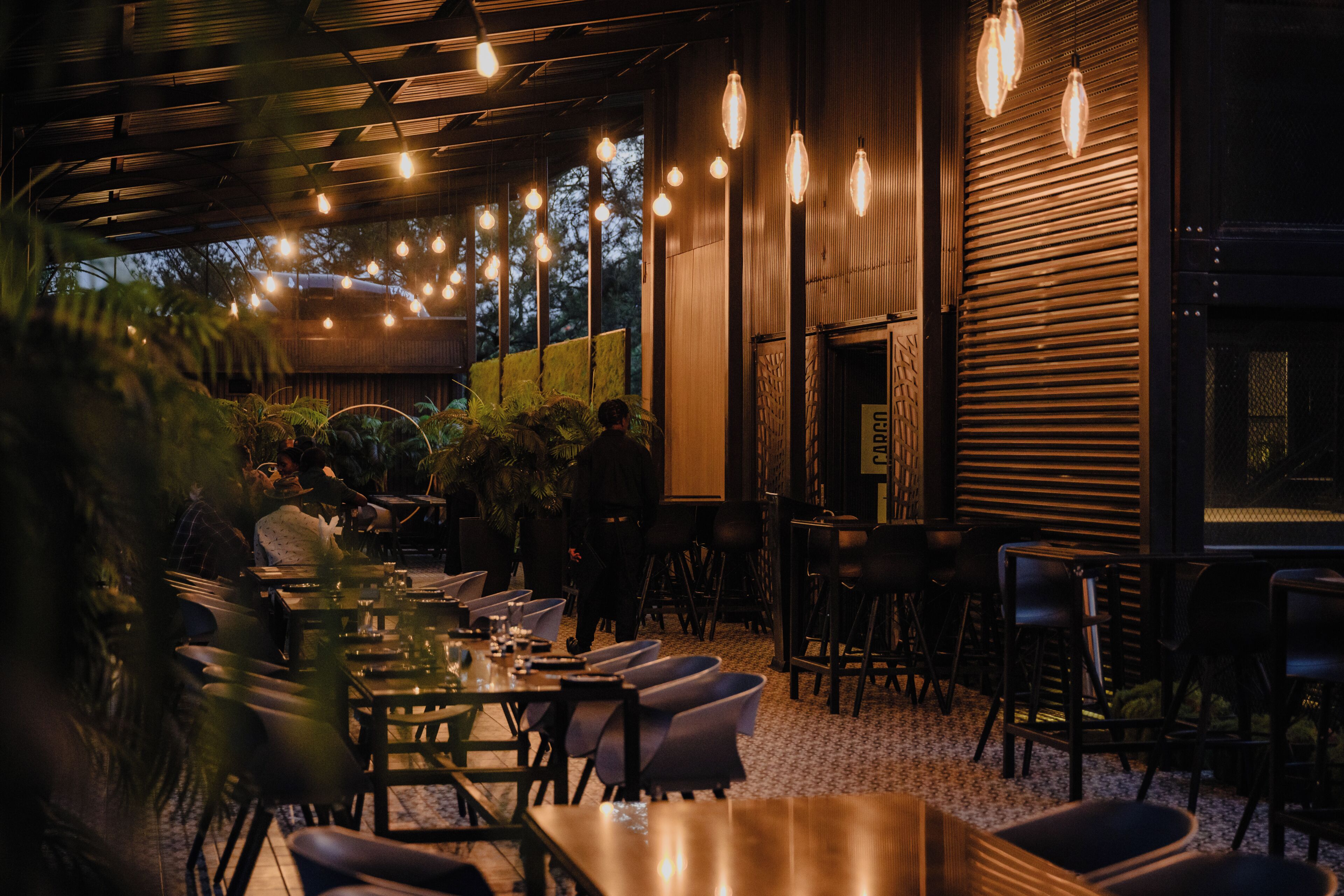 An indoor dining area with tables and chairs, hanging lights, and a wooden wall.