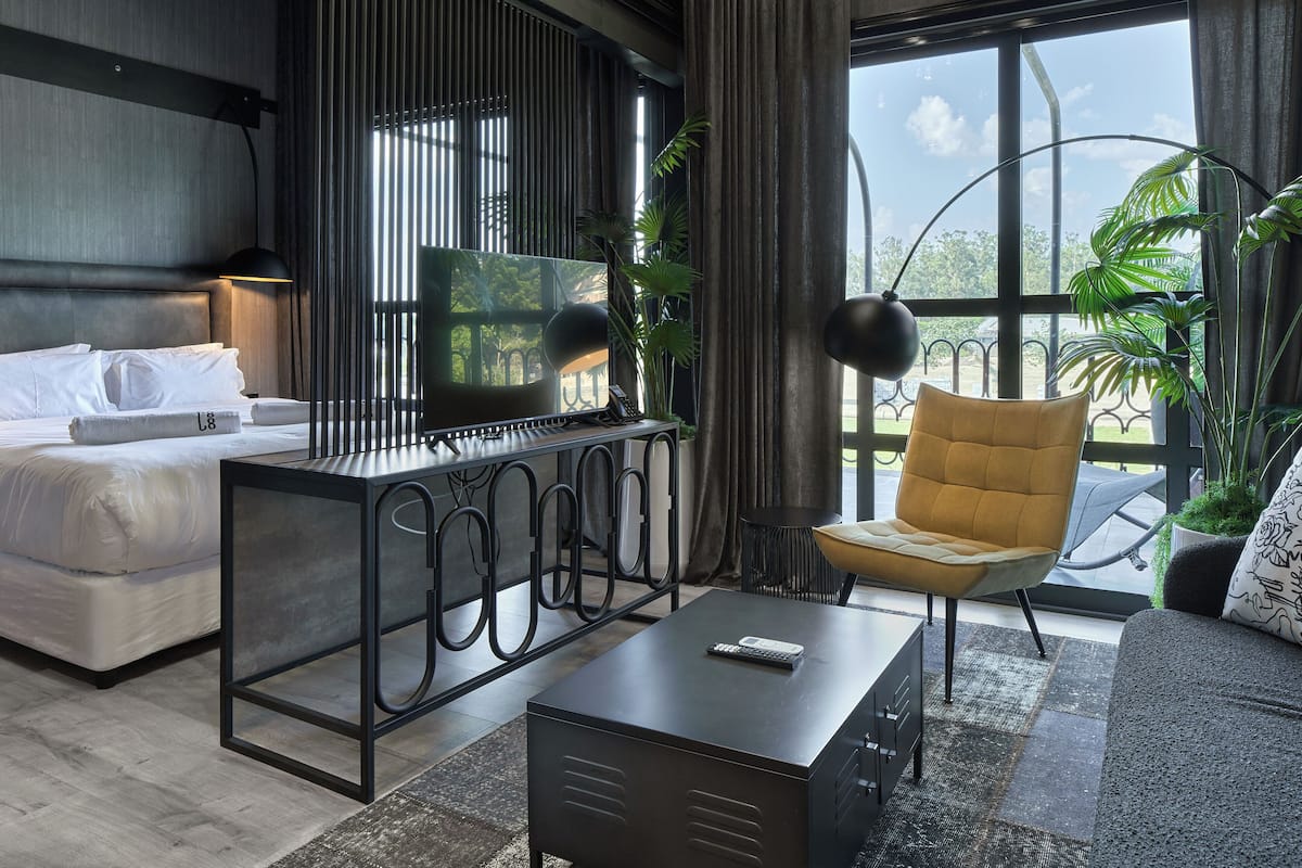 A modern hotel room with a bed, a desk, a chair, and large windows with plants.