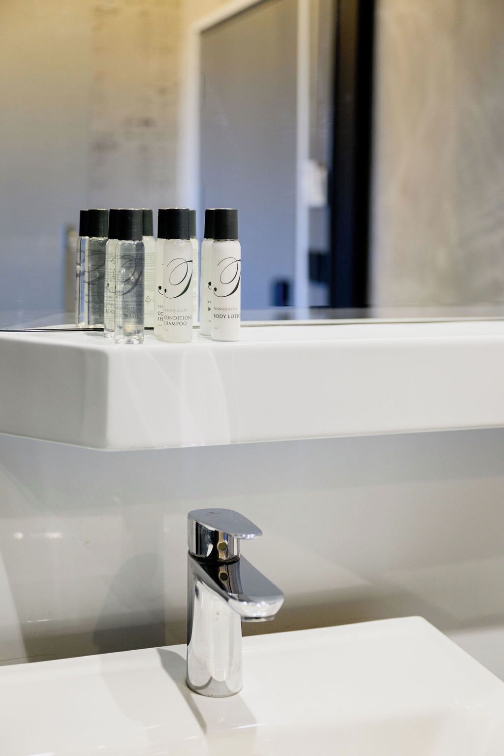 A bathroom with a white sink and a mirror above it. On the sink, there are four bottles of body lotion and shampoo.