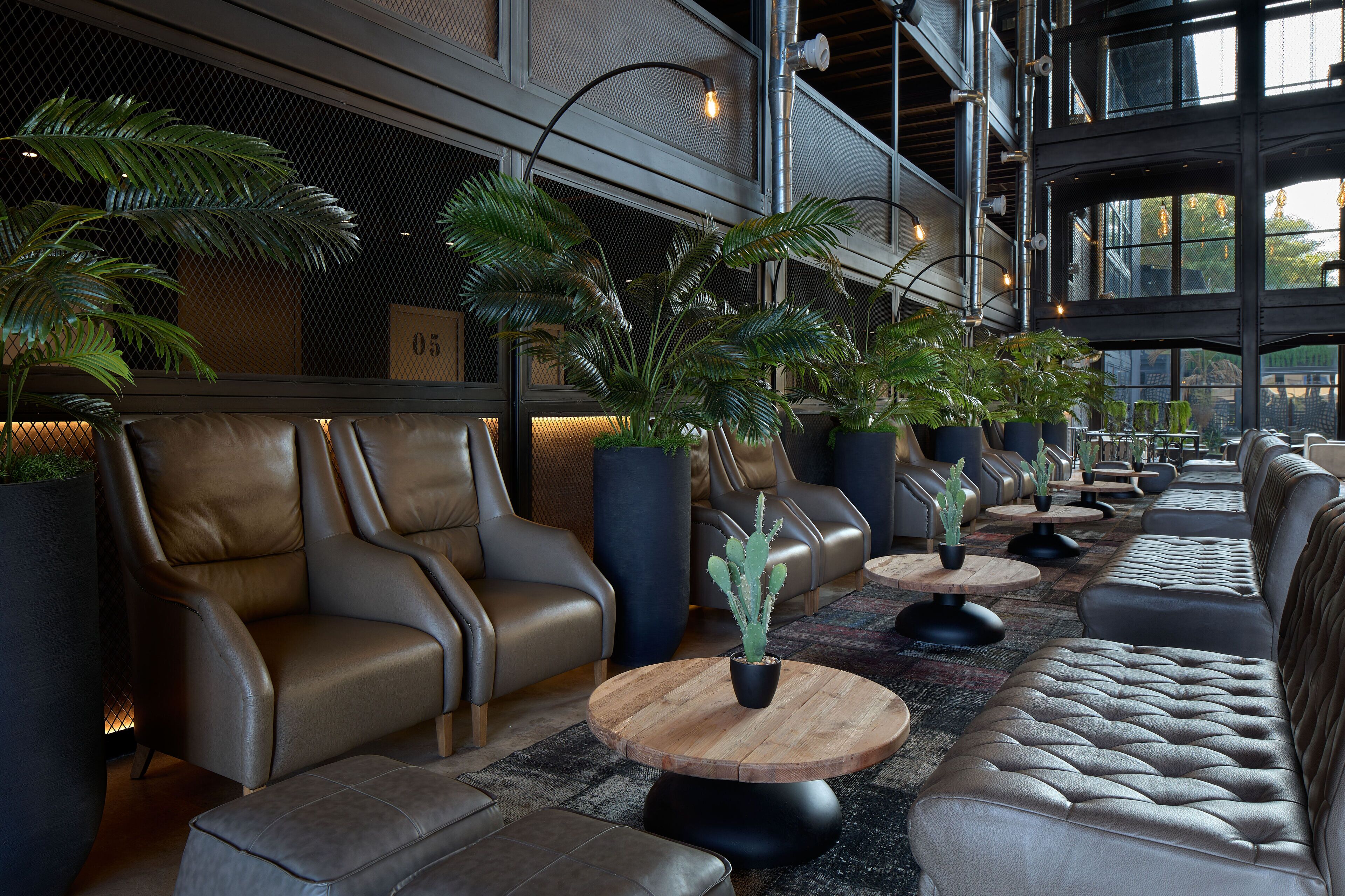 A modern lounge area with leather sofas and round wooden tables, featuring potted plants and a high ceiling with exposed beams.
