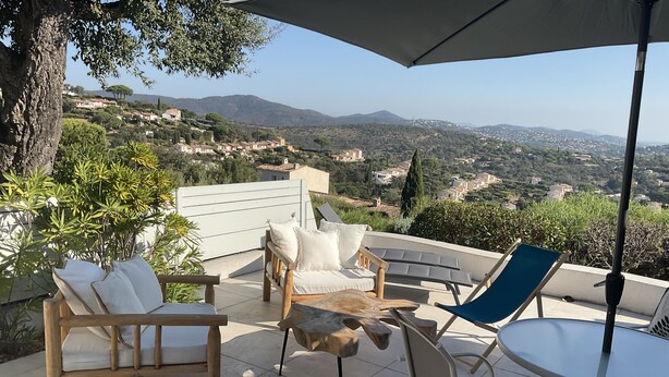 An outdoor patio with a wooden table, two white cushioned chairs, a blue chair, and an umbrella. - Xxxxxxxxxx (Sainte-Maxime)