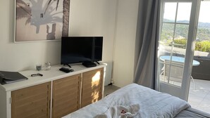 A bedroom with a bed, a TV on a wooden cabinet, a painting of palm trees, and a balcony with a table and chairs. - Xxxxxxxxxx (Sainte-Maxime)