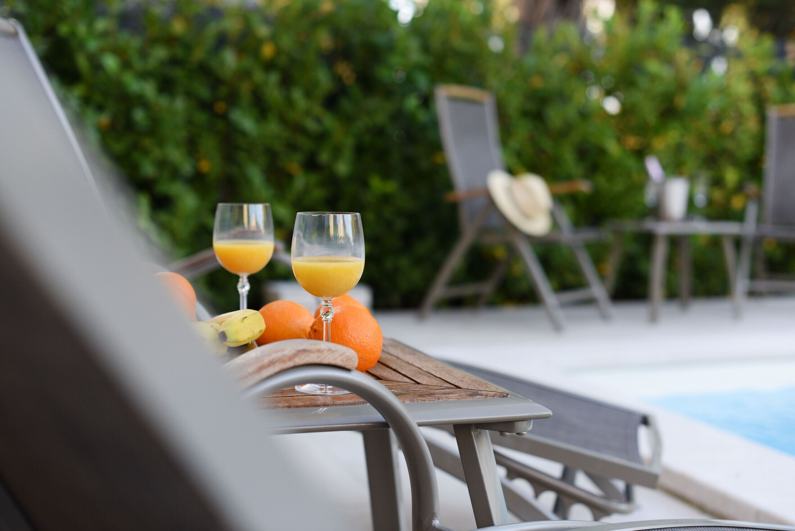 A poolside setting with lounge chairs, a wooden table with fruits and drinks, and a pool in the background.