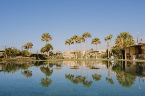 A clear pool with reflections of palm trees and buildings, surrounded by a landscaped area with a stone wall and a building with a sloped roof. - Seafront bungalow, golf, swimming pool, relax pool, 86 (San Miguel de Abona)