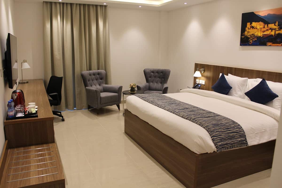 A hotel room with a large bed, two armchairs, a desk with a TV, and a painting on the wall.