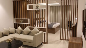 A modern hotel room with a sectional sofa, a small round table, a bed with a wooden headboard, and a wooden partition with shelves. - DUQM EXPRESS (Duqm)