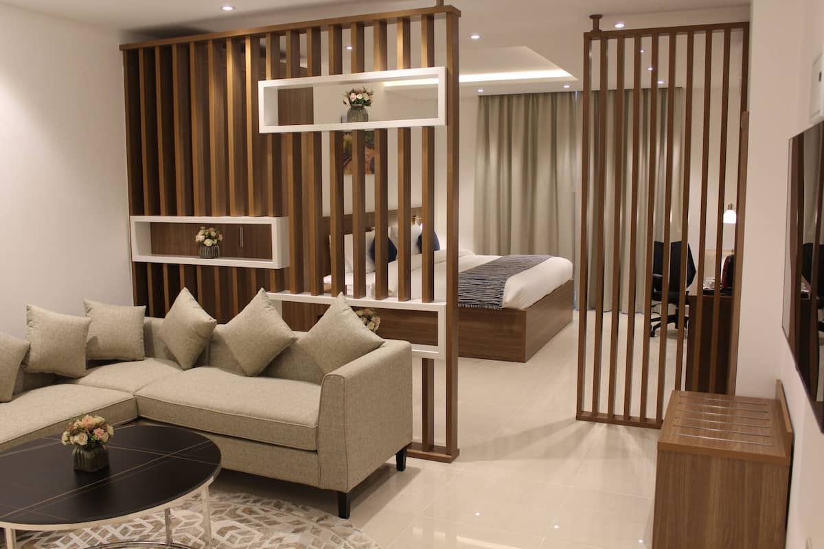 A modern hotel room with a sectional sofa, a small round table, a bed with a wooden headboard, and a wooden partition with shelves.