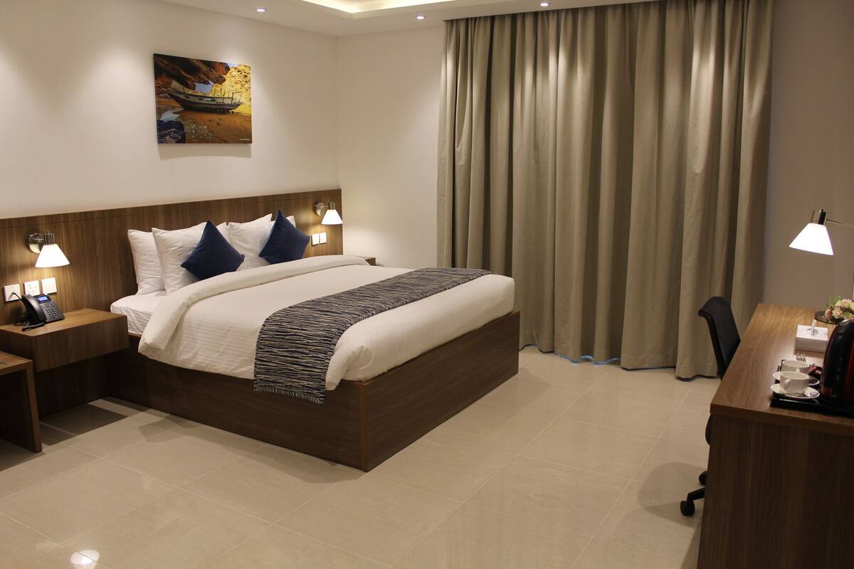 A hotel room with a large bed, a wooden headboard, two bedside lamps, a desk with a chair, and a painting on the wall.