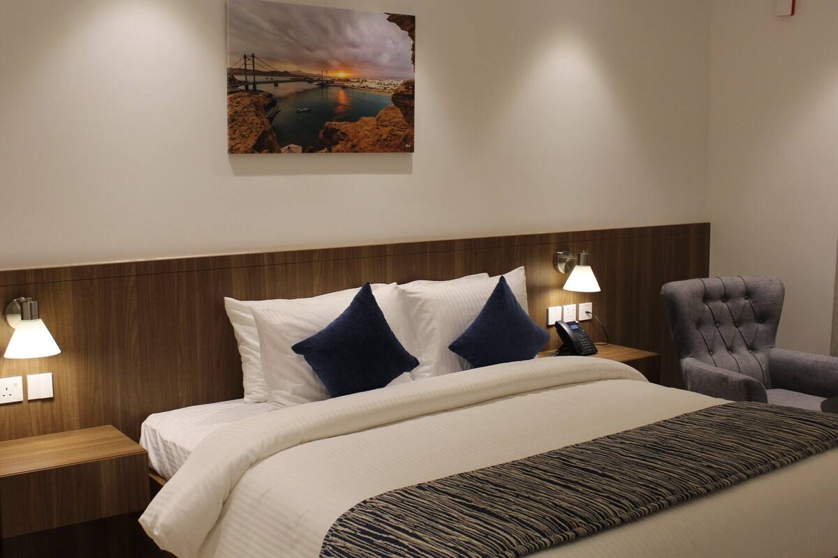 A hotel room with a bed, a nightstand, a chair, and a wall-mounted lamp.