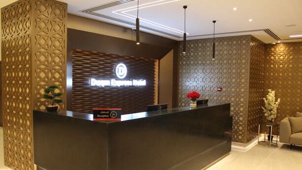 A reception desk with a modern design, a digital display, and a reception area with a sofa and decorative plants. - DUQM EXPRESS (Duqm)