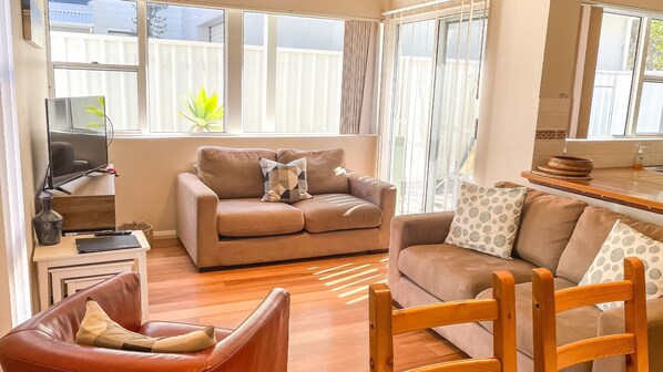A cozy living room with a sofa, armchair, and wooden coffee table. A TV is mounted on the wall, and there is a window with curtains.