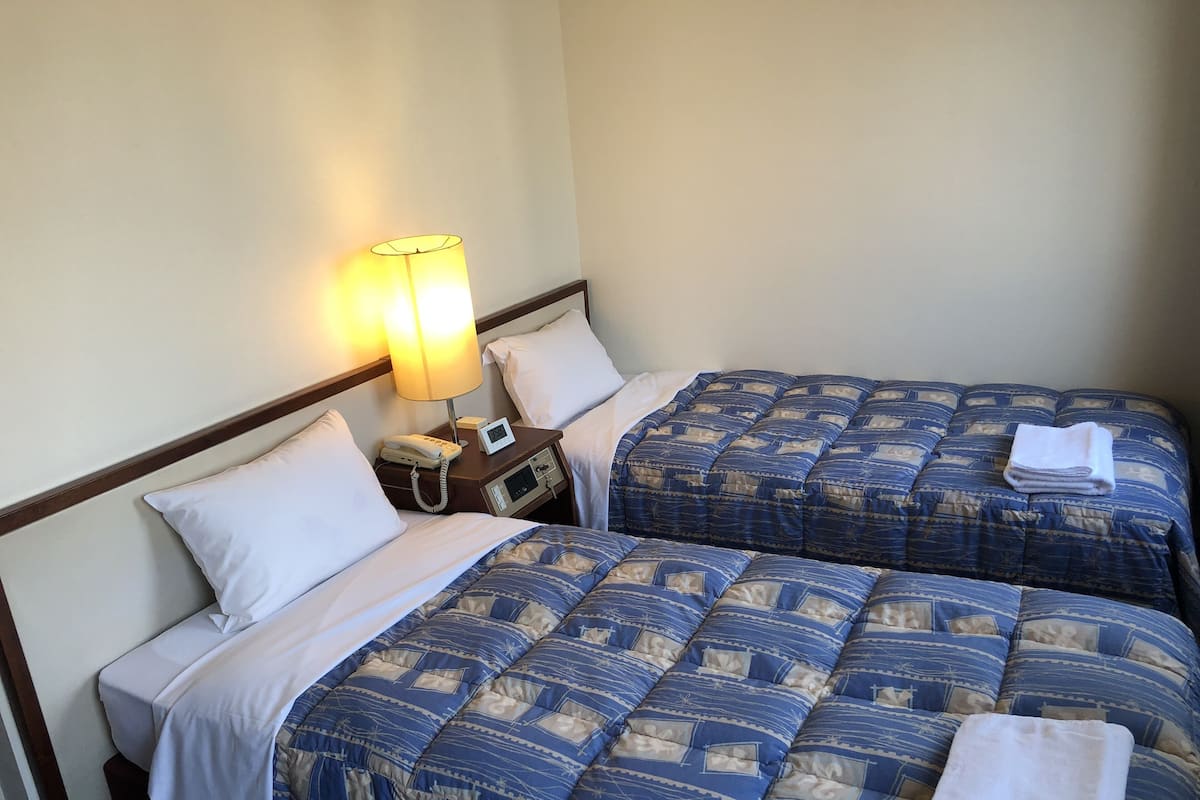 Habitación de hotel con dos camas, cada una con cubrecama azul estampado, almohadas blancas y una toalla blanca doblada sobre las camas. Hay una mesita de noche con una lámpara, un teléfono y un pequeño reloj.