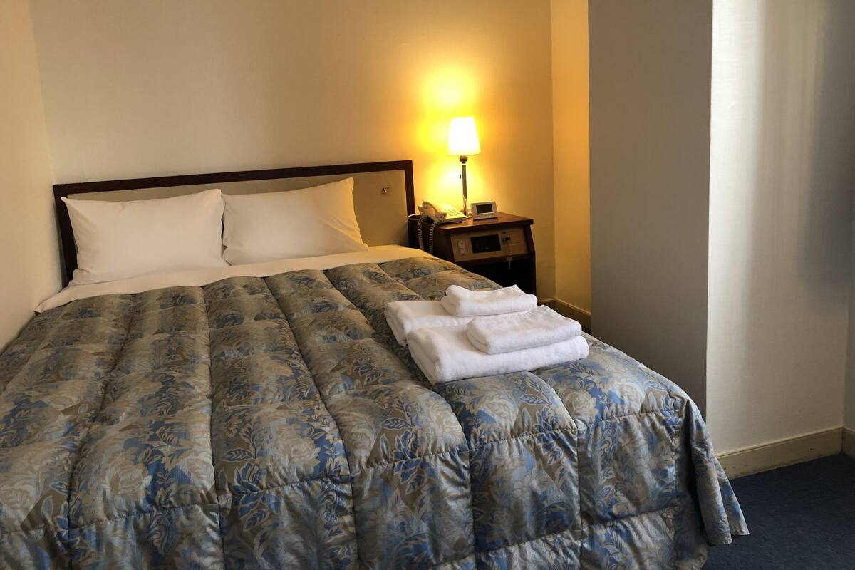 Una cama bien hecha con un edredón estampado con motivos florales, una lámpara de noche y una toalla doblada sobre la cama.
