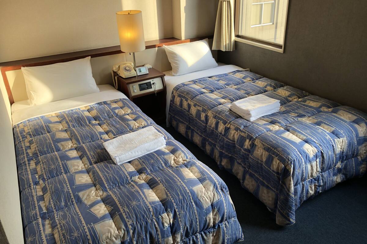 Dos camas con ropa de cama estampada en azul y beige, una mesita de noche con una lámpara y una ventana con cortinas.
