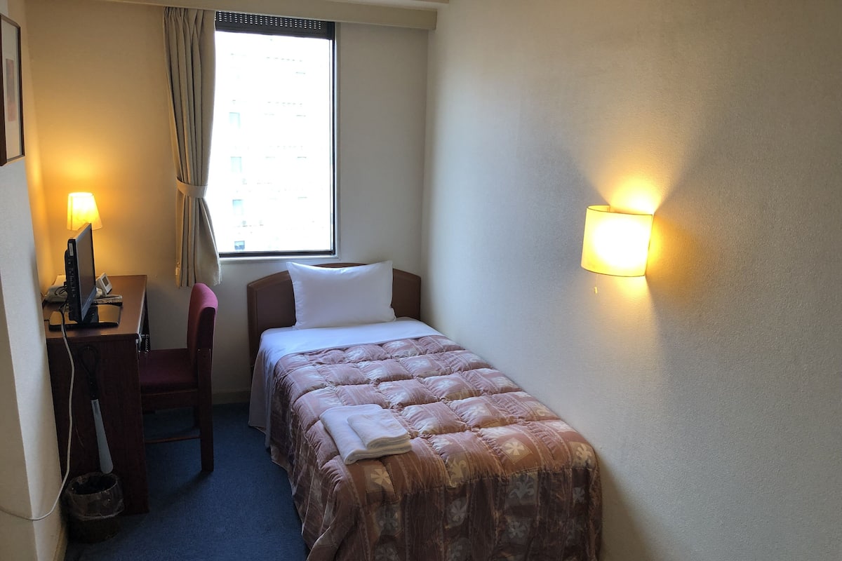 Una habitación de hotel pequeña con una cama individual, un escritorio con computadora, una silla, una ventana con cortinas y una lámpara fijada a la pared.