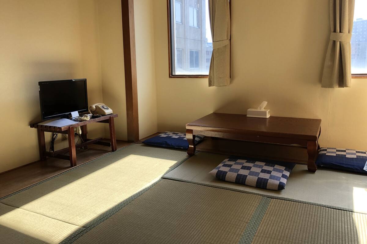 Una habitación con esteras de tatami, televisión, teléfono, mesa con caja de pañuelos y ventana con cortinas.