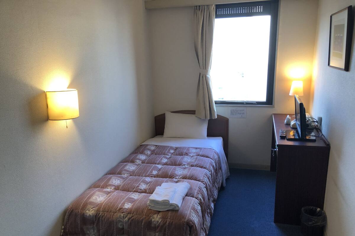 Un dormitorio pequeño y ordenado con una cama individual, un escritorio con computadora, una lámpara y una ventana con cortinas.