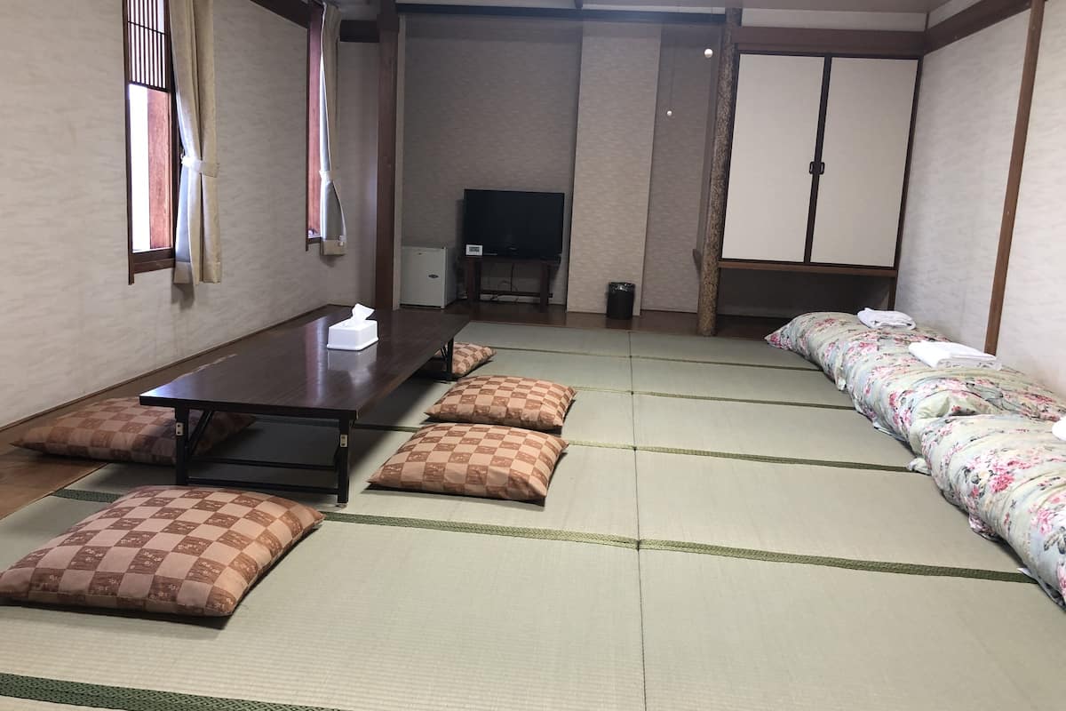 Una habitación tradicional japonesa con suelo de tatami, una mesa baja con cojines, un televisor y una cama futón.