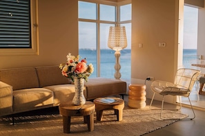 Relax in this stylish living room with a sofa, coffee table, and wicker chair, offering a view of the sea through large windows.