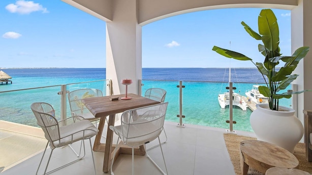 A balcony with a wooden table and chairs, a potted plant, and a view of the ocean and boats.