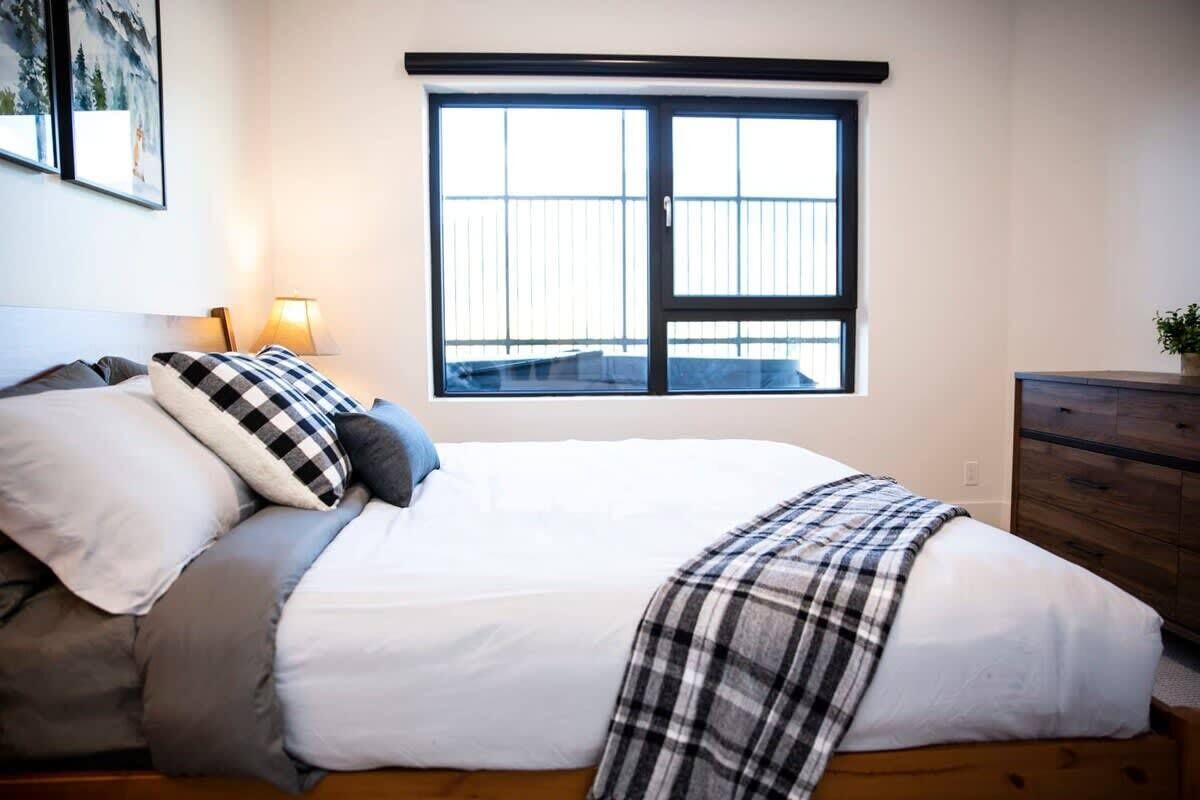 Une chambre avec une grande fenêtre, un lit avec de nombreux oreillers, une lampe de chevet et une commode.