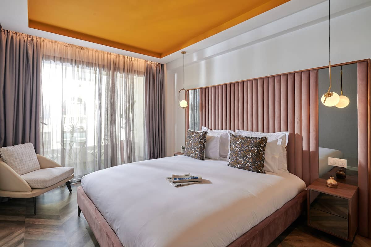 A modern hotel room with a large bed, a chair, a nightstand, and a window with curtains.