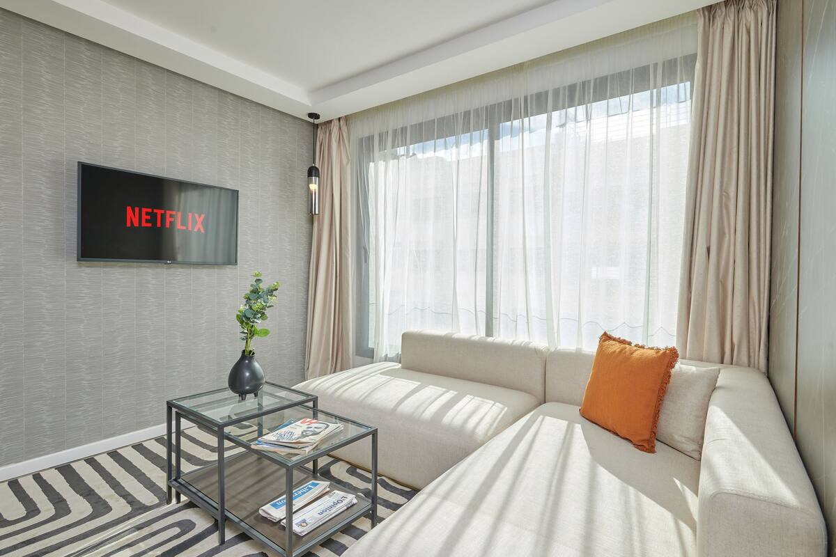 A modern living room with a sofa, a glass coffee table, a flat-screen TV displaying "NETFLIX", and sheer curtains on the windows.
