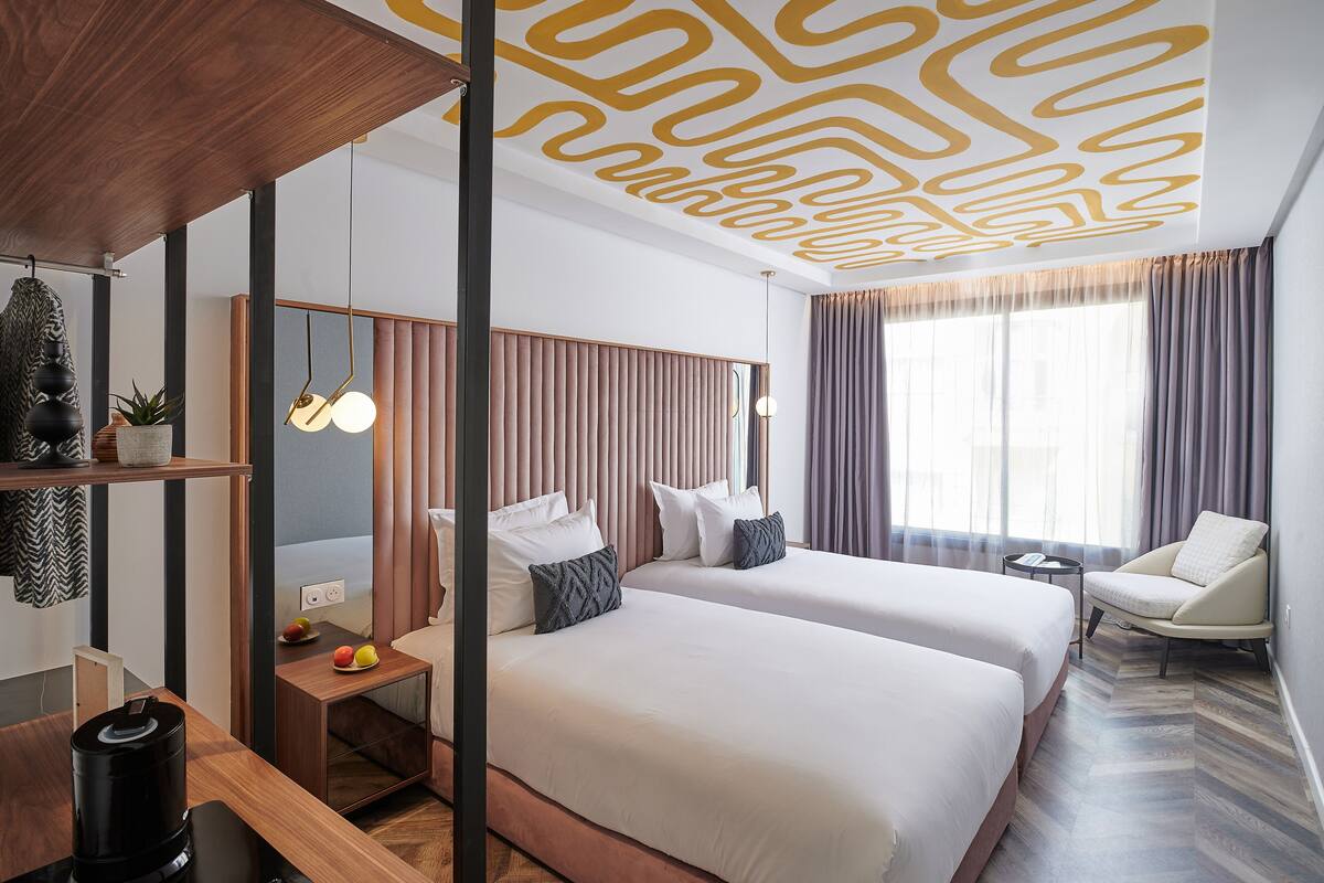A modern hotel room with two beds, a wooden desk, a chair, and a large window with curtains.