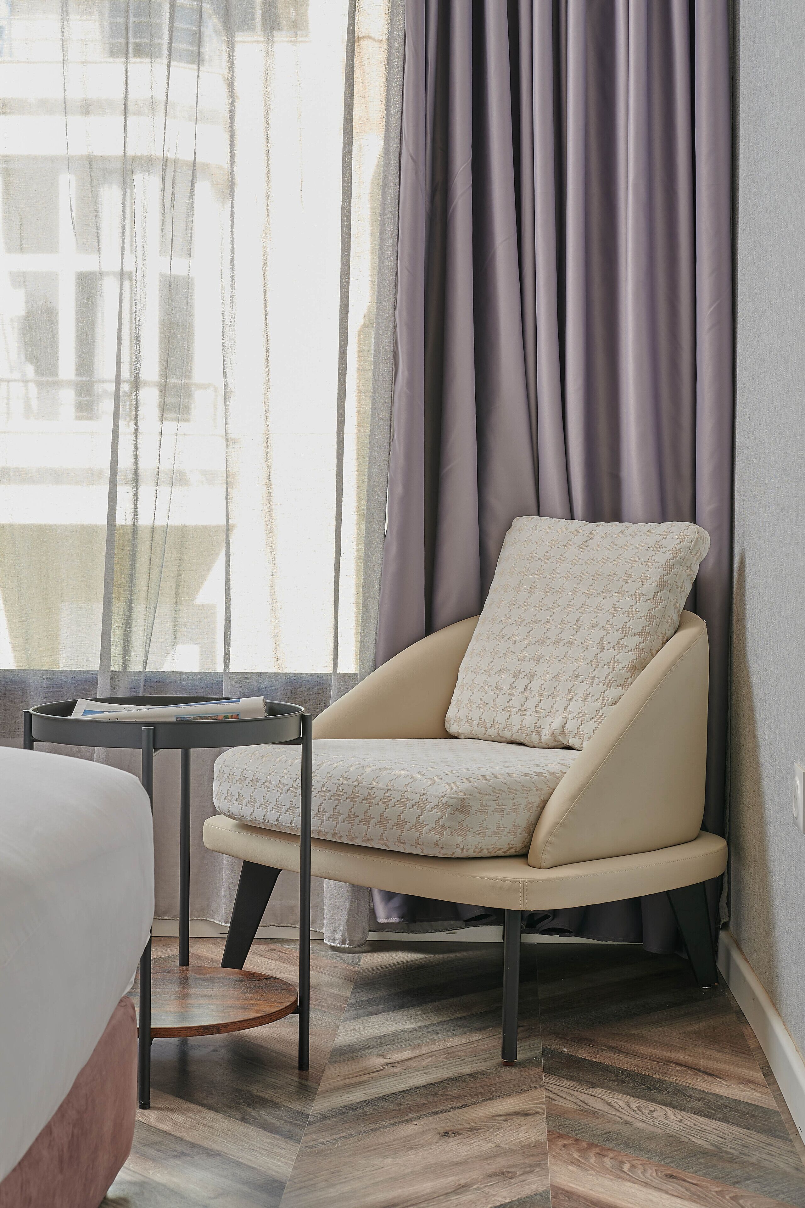 Habitación de esquina con cama, una silla con cojín, una mesita y una ventana con cortinas.